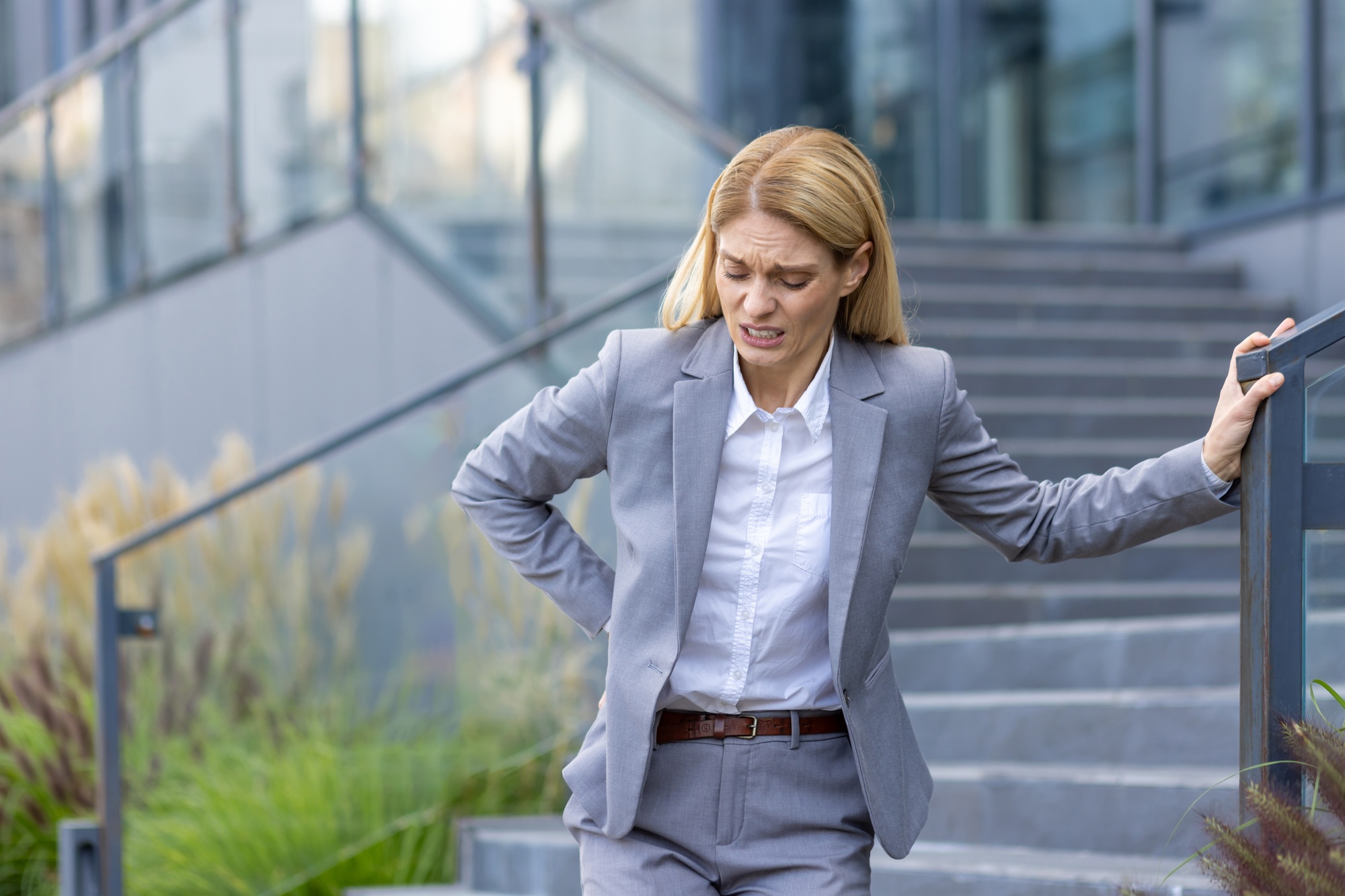 Businesswoman experiencing back pain while walking down stairs outdoors in an office setting