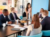 Female manager stands addressing colleagues in meeting room